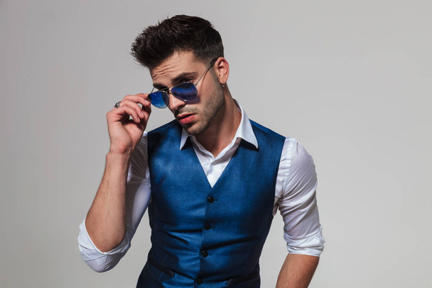 portrait of attractive man wearing a blue vest fixing sunglasses while looking to side on light grey background - Photo, Image