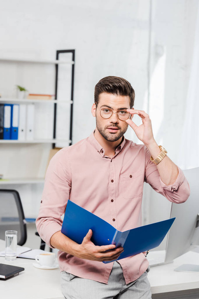 handsome young businessman with folder and eyeglasses leaning back at workplace and looking at camera - Photo, image