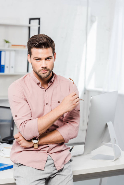 handsome young businessman leaning back at workplace and looking at camera - Fotoğraf, Görsel