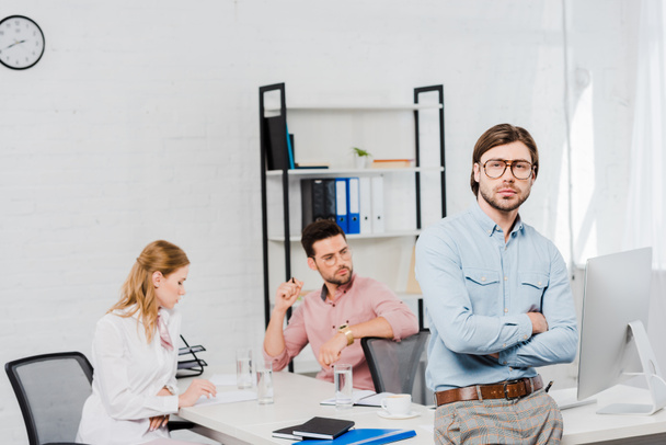 handsome young businessman with crossed arms looking at camera and leaning back on table at modern office while colleagues working together on background - Photo, Image