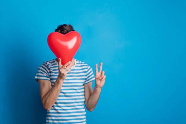 Portrait of a young man in a studio on a blue background, holding red heart. - Fotografie, Obrázek