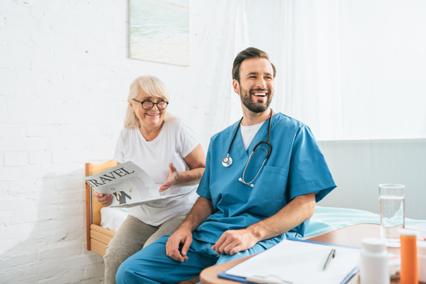 smiling young doctor with stethoscope sitting on hospital bed with happy senior woman reading travel newspaper - Photo, Image