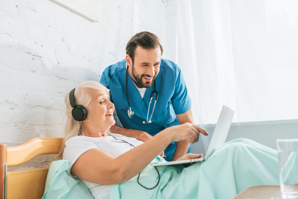 Lächelnder Krankenpfleger blickt glückliche Seniorin mit Kopfhörer an und benutzt Laptop im Krankenhausbett    - Foto, Bild
