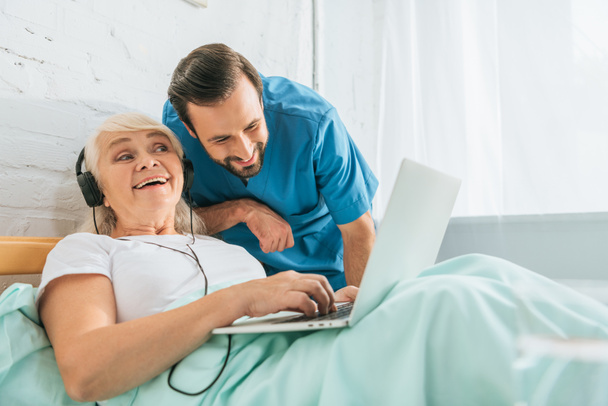 病院のベッドでノート パソコンを使用してヘッドフォンで幸せの年配の女性を見て笑みを浮かべて医師    - 写真・画像