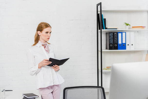 serious young businesswoman with book standing at modern office and looking away - Photo, Image