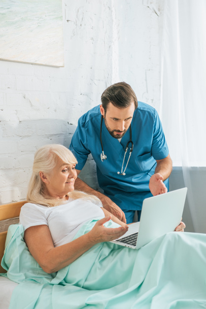 молодий лікар зі стетоскопом дивиться на старшу жінку, використовуючи ноутбук у лікарняному ліжку
  - Фото, зображення
