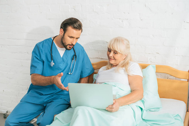 enfermeiro com estetoscópio olhando para a mulher idosa usando laptop na cama do hospital
 - Foto, Imagem