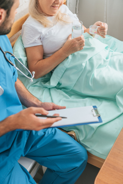 cropped shot of doctor writing on clipboard while senior woman taking medicine in bed - Foto, Imagen