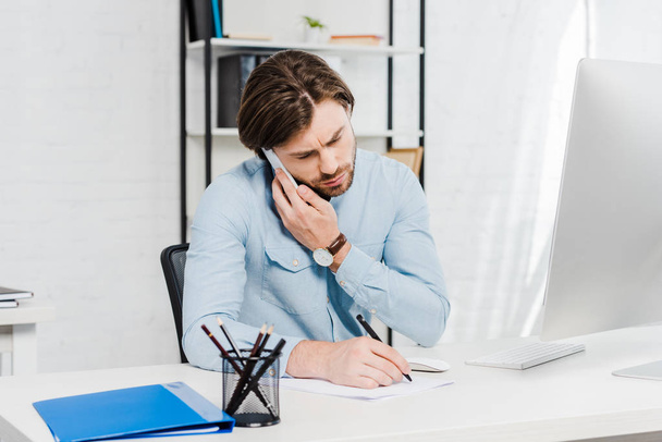 confident young businessman talking by phone and making notes at workplace - Photo, image