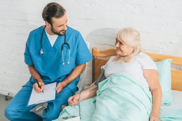 high angle view of male nurse writing on clipboard while measuring blood pressure to smiling senior woman lying in bed - Photo, Image