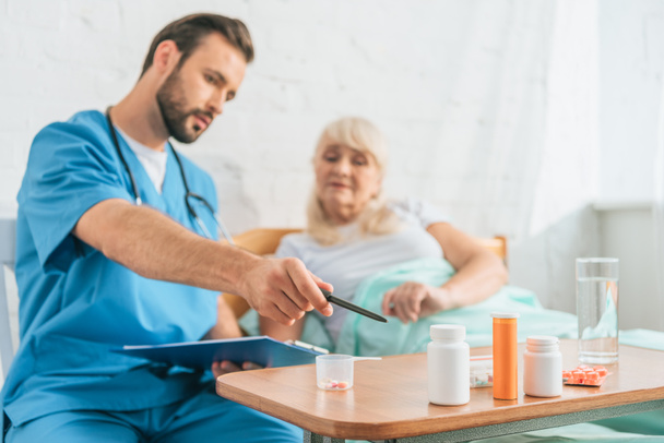 Krankenpfleger hält Klemmbrett und zeigt auf Tabletten auf Seniorin, die im Bett liegt - Foto, Bild
