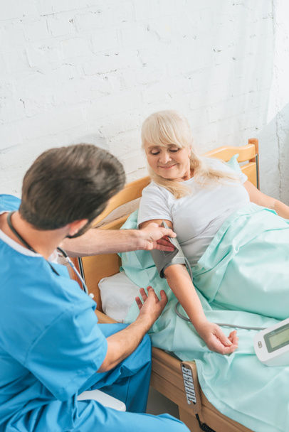 mladý doktor měření krevního tlaku a starší žena v nemocniční posteli - Fotografie, Obrázek