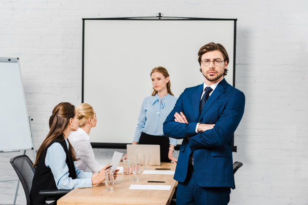 handsome young businessman with crossed arms looking at camera at modern office with female colleagues sitting on background - 写真・画像