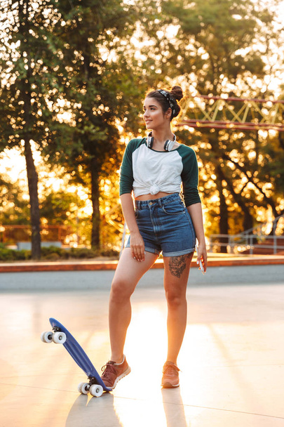 Cheerful young girl with headphones and skateboard standing at the park - Fotoğraf, Görsel