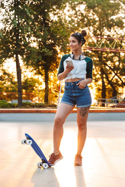 Cheerful young girl with headphones and skateboard standing at the park and using mobile phone - Zdjęcie, obraz
