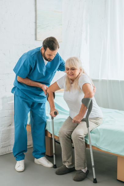 чоловіча медсестра допомагає старшій жінці з милицями, сидячи на лікарняному ліжку
  - Фото, зображення