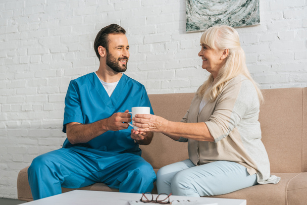 sorrindo enfermeira do sexo masculino dando copo com bebida quente para a mulher idosa feliz
 - Foto, Imagem