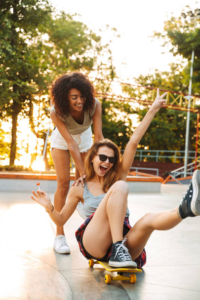 Two happy young girls having fun while riding on a skateboard at the park - Фото, изображение