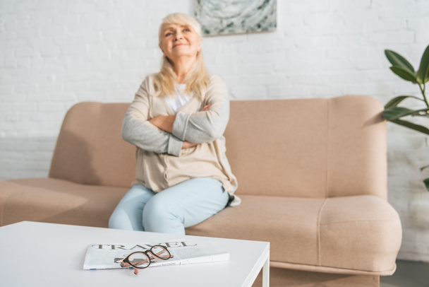 travel newspaper and eyeglasses on table and smiling senior woman sitting on couch behind - Photo, image