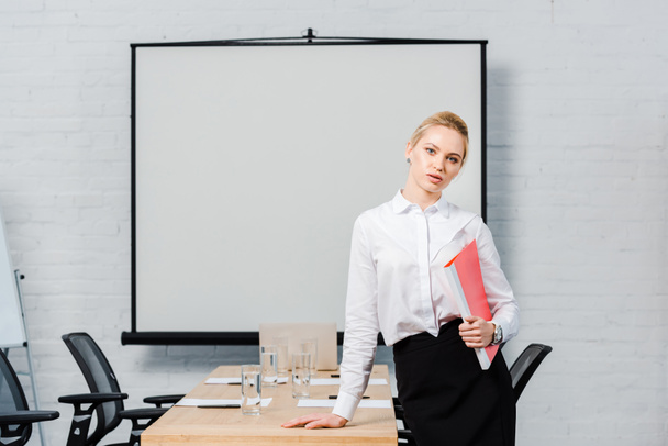 attractive young businesswoman with folder looking at camera in conference hall with blank presentation board - Φωτογραφία, εικόνα