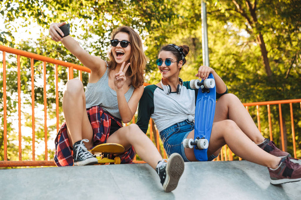 Two smiling young girls having fun while sitting with skateboards at the park and taking selfie - Фото, изображение