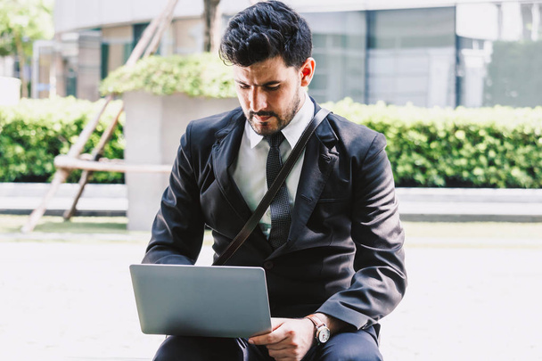 Przystojny biznesmen w czarny elegancki garnitur siedzi i praca z laptopem na miasto. Biznes i uruchamiania pomysłem - Zdjęcie, obraz