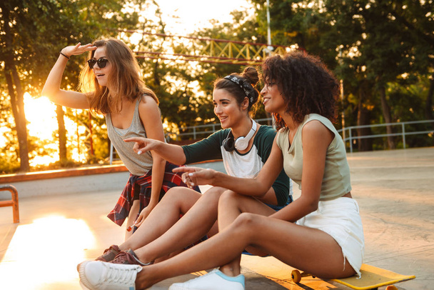 Image of young happy three friends woman outdoors with skateboard sitting talking with each other looking aside. - Φωτογραφία, εικόνα