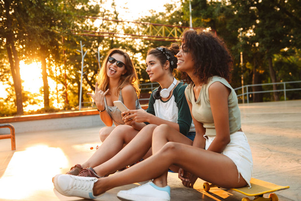 Image of young happy three friends woman outdoors with skateboard sitting talking with each other using mobile phone. - Zdjęcie, obraz