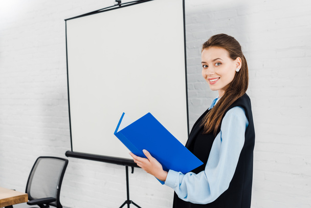 smiling young businesswoman with folder of documents looking at camera in front of blank presentation board - Foto, immagini