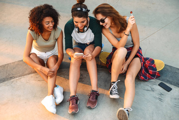 Image of young happy three friends woman outdoors sitting talking with each other using mobile phones listening music with earphones - Zdjęcie, obraz