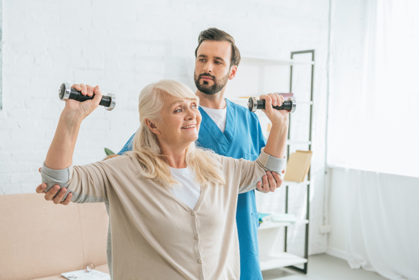 social worker looking at smiling senior woman training with dumbbells  - Photo, Image
