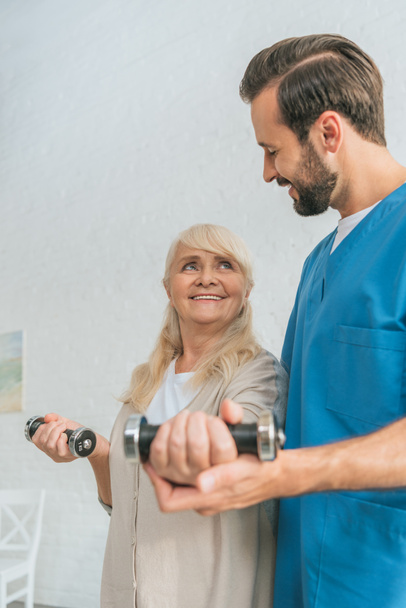 smiling social worker helping happy senior woman exercising with dumbbells - Foto, Bild