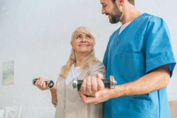 cropped shot of social worker helping smiling senior woman exercising with dumbbells - Foto, Bild