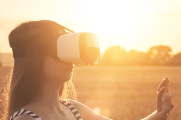 Belle fille dans un champ de blé doré utilise des lunettes de réalité virtuelle au coucher du soleil - Photo, image