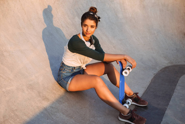 Pretty young girl with headphones and skateboard sitting at the ramp - Foto, Imagen