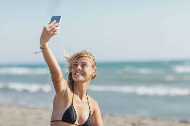 Pretty Caucasian woman sitting on sandy beach and taking selfie with her smartphone. - Photo, image