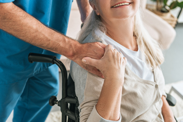 cropped shot of smiling senior woman in wheelchair holding hand of caregiver - Фото, изображение