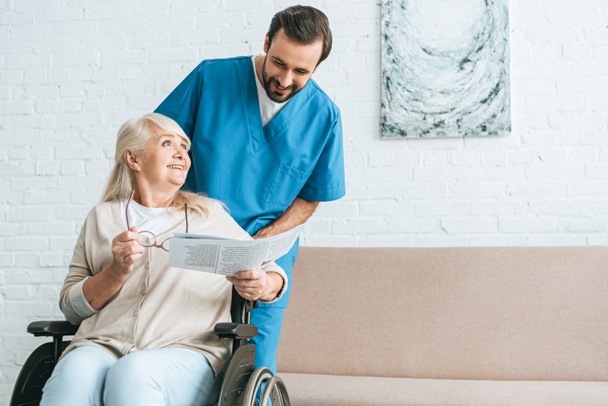 Lächelnde junge Krankenschwester blickt fröhliche Seniorin an, die im Rollstuhl Zeitung liest  - Foto, Bild
