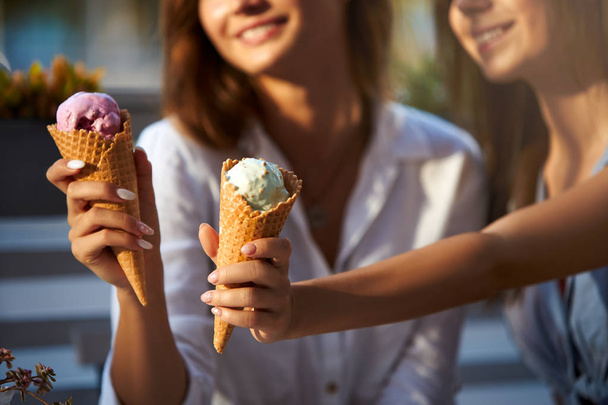 Close up shot of ice cream cones in hand of a woman standing with her friend. Two young women outdoors eating icecream on a sunny day. Isolated view, no face, copyspace for designers. Summer theme. - Φωτογραφία, εικόνα