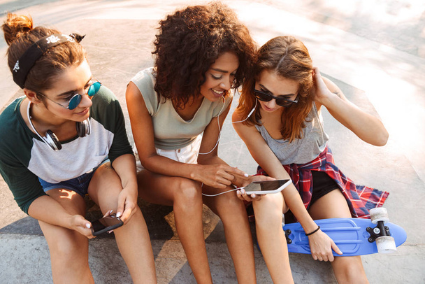 Imagen de la joven feliz mujer tres amigos al aire libre sentado hablando entre sí usando teléfonos móviles escuchando música con auriculares
. - Foto, imagen