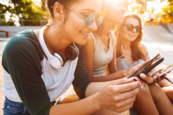 Image of young happy three friends woman outdoors sitting talking with each other using mobile phones listening music with earphones. - Fotó, kép