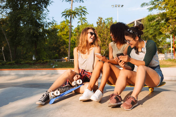 Image of young happy three friends woman outdoors sitting talking with each other using mobile phones listening music with earphones. - Photo, Image