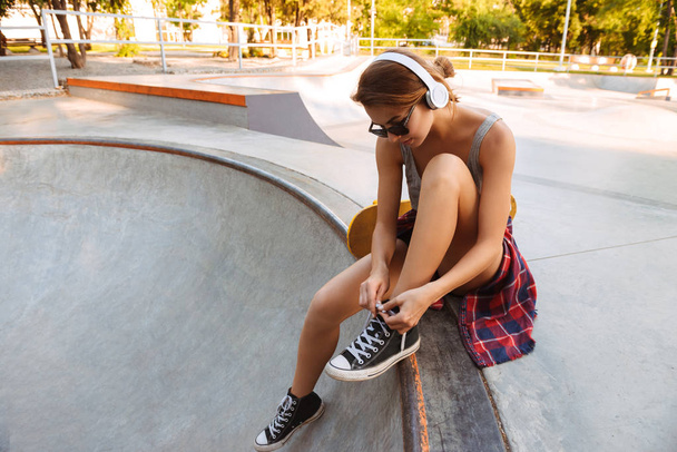 Pretty young girl in headphones listening to music while sitting with skateboard at a park and tying shoelaces - Фото, изображение