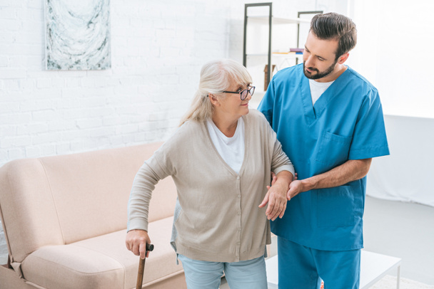 male nurse supporting senior woman in eyeglasses with walking stick  - Photo, Image