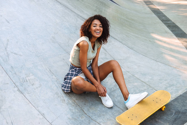 Laughing young afro american girl sitting with skateboard at a park - Fotoğraf, Görsel