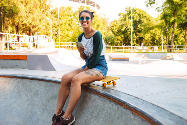 Photo of young happy cheerful woman sitting outdoors with skateboard using mobile phone laughing. - Φωτογραφία, εικόνα