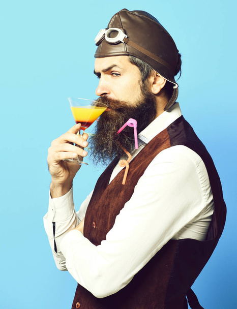 holding glass of alcoholic beverage in vintage suede leather waistcoat with hat and glasses on red studio background. serious handsome bearded pilot - Фото, зображення