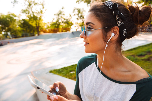 Photo of young smiling pretty woman outdoors listening music with earphones using mobile phone. - Valokuva, kuva