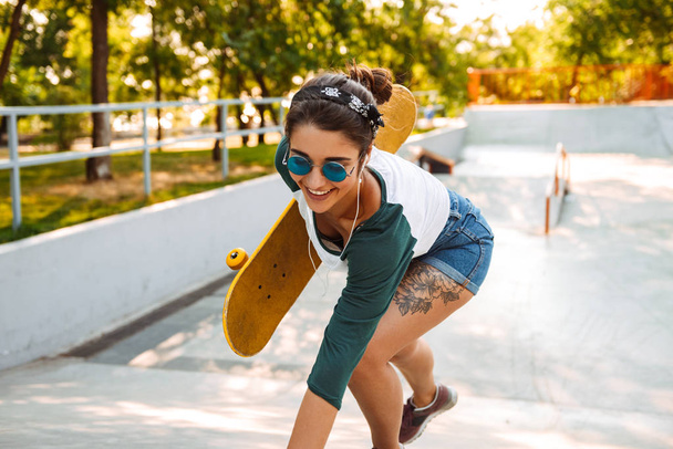 Cheerful young girl in eaephones riding on a skateboard at a park - Фото, изображение