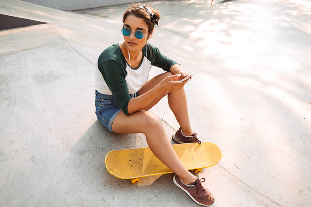 Pretty young girl in earphones listening to music while sitting with skateboard at a park - 写真・画像